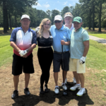 Group of five adults posing for a photo on a sunny golf course, with trees in the background.
