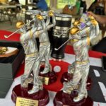 Three silver and gold-toned golf trophy statues mid-swing on maroon bases, plaques read '1st PLACE 2026 Rumble in the Morning Who's Your Caddy Summer Edition' with a blurred outdoor gathering in the background.
