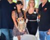 Four adults posing under a wooden pavilion; a woman in a leopard-print dress holds a plush owl with others smiling nearby.