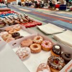 Box of assorted donuts and pastries on a table at a large indoor donation drive, with volunteers and equipment set up in the background.