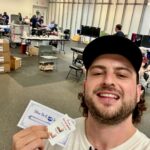 Smiling man in a brimmed cap taking a selfie at a donation event, holding Blue Bell gift cards in front of a busy room with tables and people in the background.