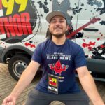 Smiling man in a blue t‑shirt and cap poses in a wide stance in front of a promotional van with 99 branding; wearing a Blood Drive badge labeled Nick.