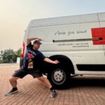 Man striking a wide lunge pose beside a white blood-donation van that says “Please give blood.”