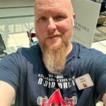 Man with a bald head and long beard wearing a navy T‑shirt featuring a cartoon blood drop lifting weights; mall interior in background, name badge reads Sean Hood.