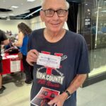 Smiling older man with glasses holding a Blue Bell Ice Cream ticket and a guitar brochure at an indoor event.