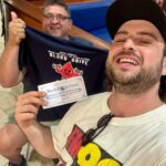Two men pose for a selfie in a mall; one holds a dark T‑shirt with a blood drive logo and a certificate that says Blue Bell Ice Cream, smiling.