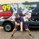Man in a cap and blue shirt striking a martial-arts pose beside a FM99.com promotional van.