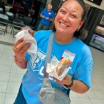 Smiling woman in a blue T-shirt holding two donuts in napkins inside a shopping mall; storefront sign behind reads 'CHOI's WATCH & JEWELRY REPAIR'.