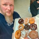 Man with a long beard takes a selfie while holding an open box of assorted donuts in a store or mall.