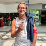 Smiling man with glasses in a white tank top holds a small snack in a shopping mall, jacket over his shoulder and a red armband on his arm.