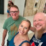 Three people pose for a selfie in a mall: smiling woman in a blue top between two men, one with glasses and a beard, all wearing name badges.