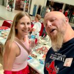 Smiling woman in a pink top poses for a selfie with a bearded man at a candy fundraising table in a mall, storefronts in the background.