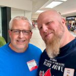 Two men pose for a selfie in a mall; one wears glasses and a blue shirt with a name badge, the other has a long beard and a graphic tee with a red cartoon character lifting weights in the foreground.