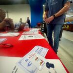 Person in a blue T-shirt stands at a red information table with brochures and cards, others talking in the background at an indoor event.