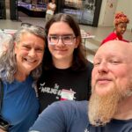 Three adults taking a selfie in a mall; woman in blue, young man with glasses, bearded man in a navy shirt, with storefronts and a shopper in the background.