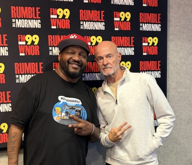 Two men pose for a photo in front of a 'Rumble in the Morning' backdrop; left man in a black shirt and red cap, right man in a light gray zip sweater, both flashing peace signs.