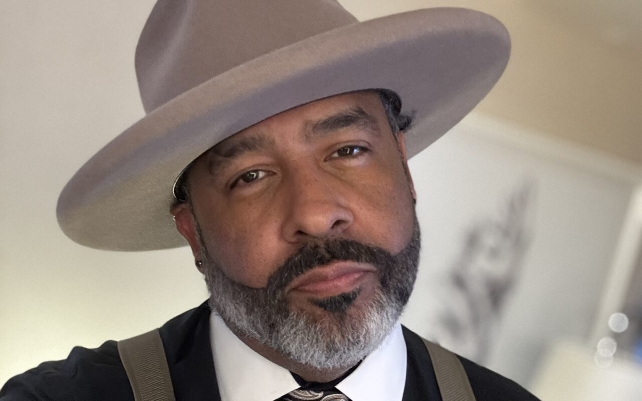 Portrait of a man with a gray beard wearing a wide-brim beige hat, white shirt, tie, and suspenders indoors.