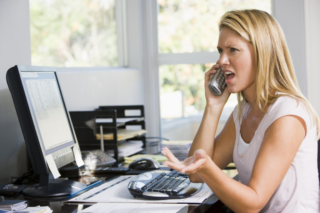 Woman at a cluttered office desk, talking on a corded phone and gesturing with her other hand.