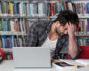 Young man in a library studies at a laptop, head in hand, surrounded by books on shelves and table.