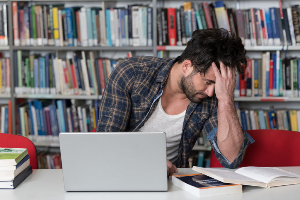 Young man in a library studies at a laptop, head in hand, surrounded by books on shelves and table.