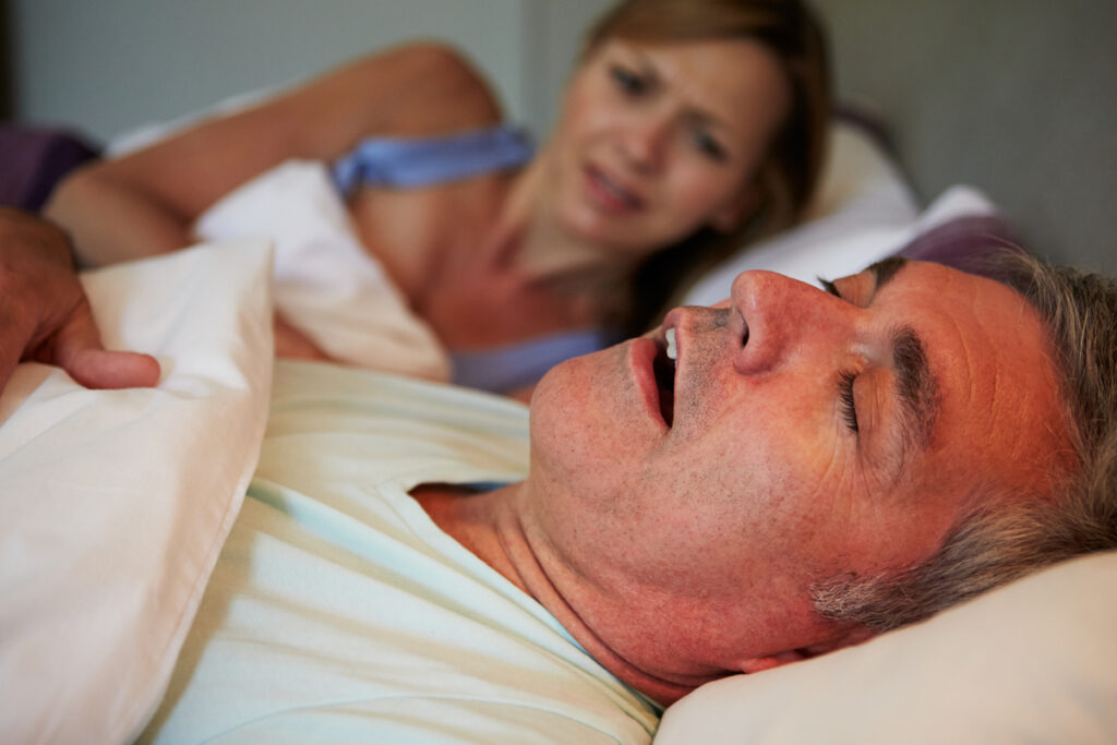 Man sleeping in bed with his head on a pillow; a concerned woman sits in the background.