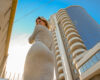 Woman in a light gray dress stands beside a tall modern building with rounded balconies and a glass tower against a blue sky, looking over her shoulder.
