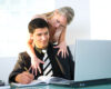 Man in a dark suit writes in an open notebook at a desk while a woman leans over his shoulder with arms around him, looking at his laptop.