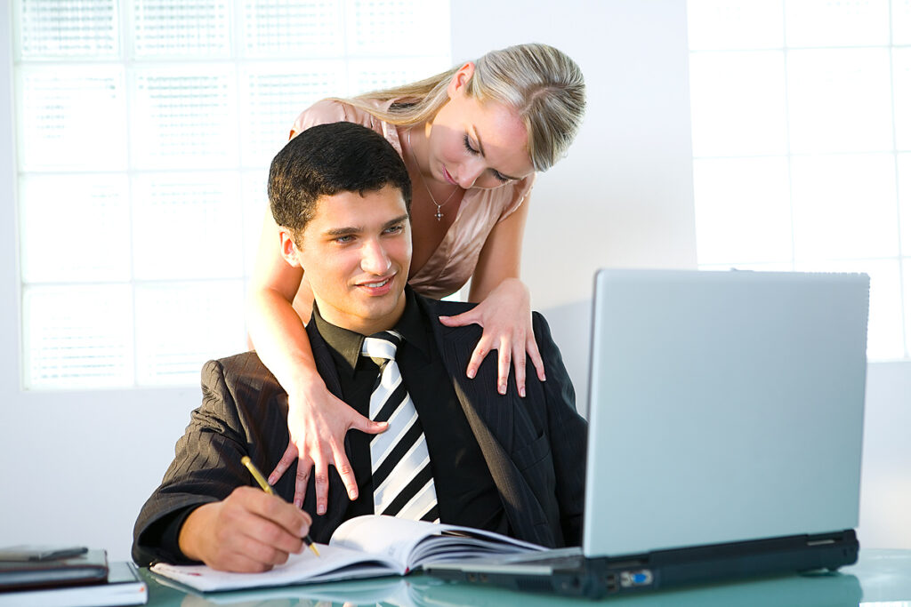 Man in a dark suit writes in an open notebook at a desk while a woman leans over his shoulder with arms around him, looking at his laptop.