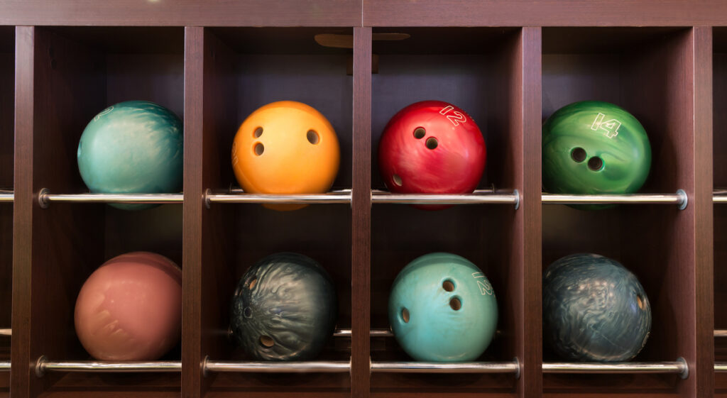 Colorful bowling balls stored in wooden cubbies on metal racks (orange, red, green, teal, pink).