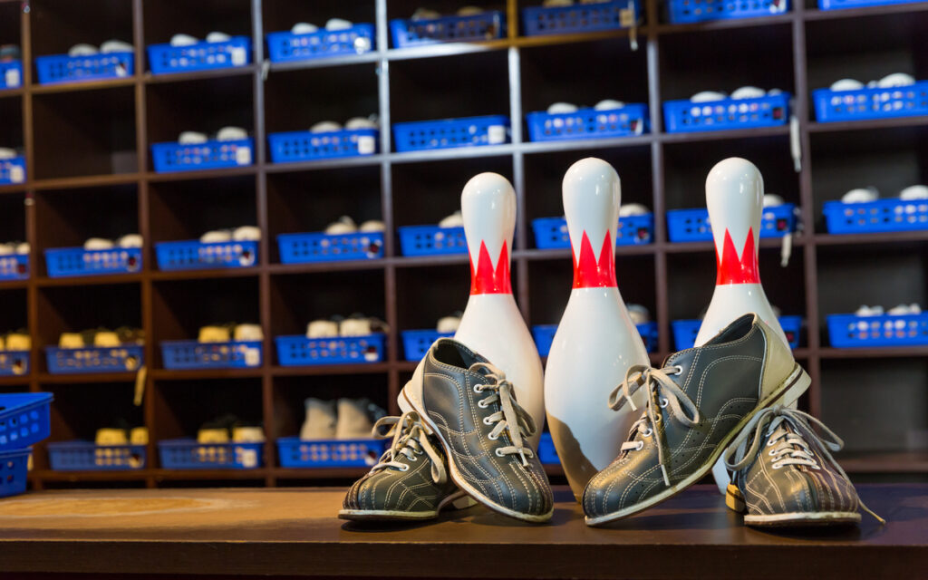 Three bowling pins with red stripes stand between two pairs of gray bowling shoes on a wooden lane.
