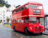 Red double-decker heritage bus parked on a coastal street with white Victorian buildings in the background.