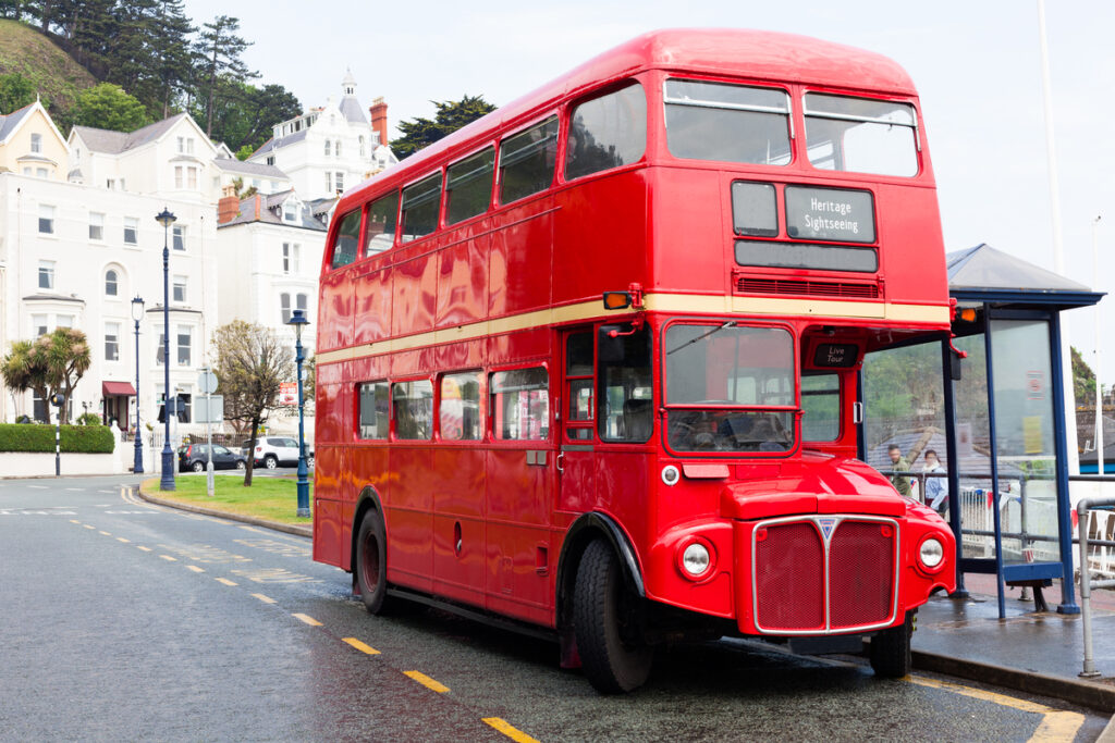 Red double-decker heritage bus parked on a coastal street with white Victorian buildings in the background.