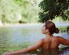 Woman with her back to the camera relaxing in a calm lake, surrounded by green trees.