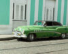 Vintage green classic car with a black roof parked on a cobblestone street in front of a turquoise building.