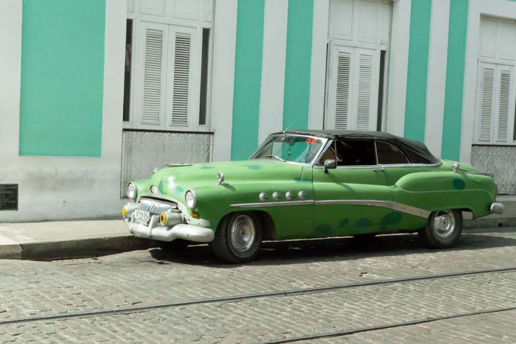 Vintage green classic car with a black roof parked on a cobblestone street in front of a turquoise building.