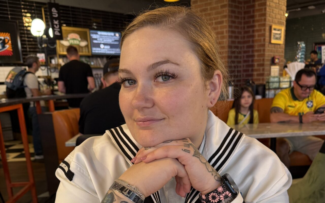 Young woman with colorful tattoos sits at a cafe table, hands folded under her chin and a subtle smile, facing the camera.