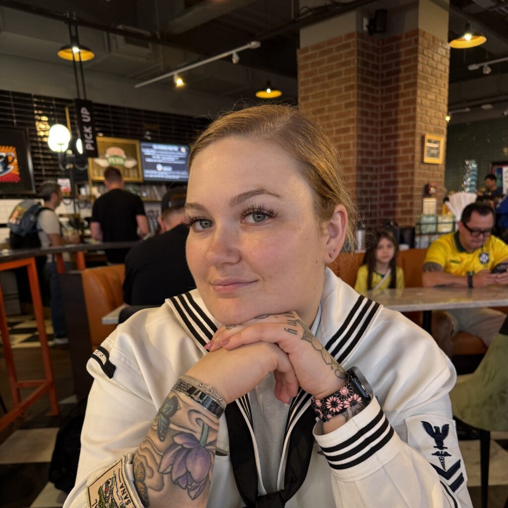 Young woman with colorful tattoos sits at a cafe table, hands folded under her chin and a subtle smile, facing the camera.