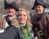 Three people in pirate costumes pose for a selfie outdoors, with a woman center and two men in tricorn hats beside her.