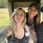 Three friends posing for a selfie in a golf cart on a sunny grassy field with trees in the background, all smiling.