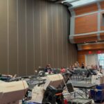 Large room with high ceiling and curved wall panels; several chairs and medical machines set up for a blood donation or medical event, with people seated and volunteers moving around.