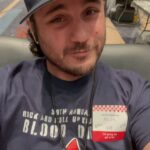 Man at a blood drive wearing a dark cap, donor badge, and a shirt with a cartoon blood drop.