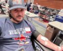 Man sitting in a donation chair, donating blood with a needle in his arm and bandage, at a blood drive center.