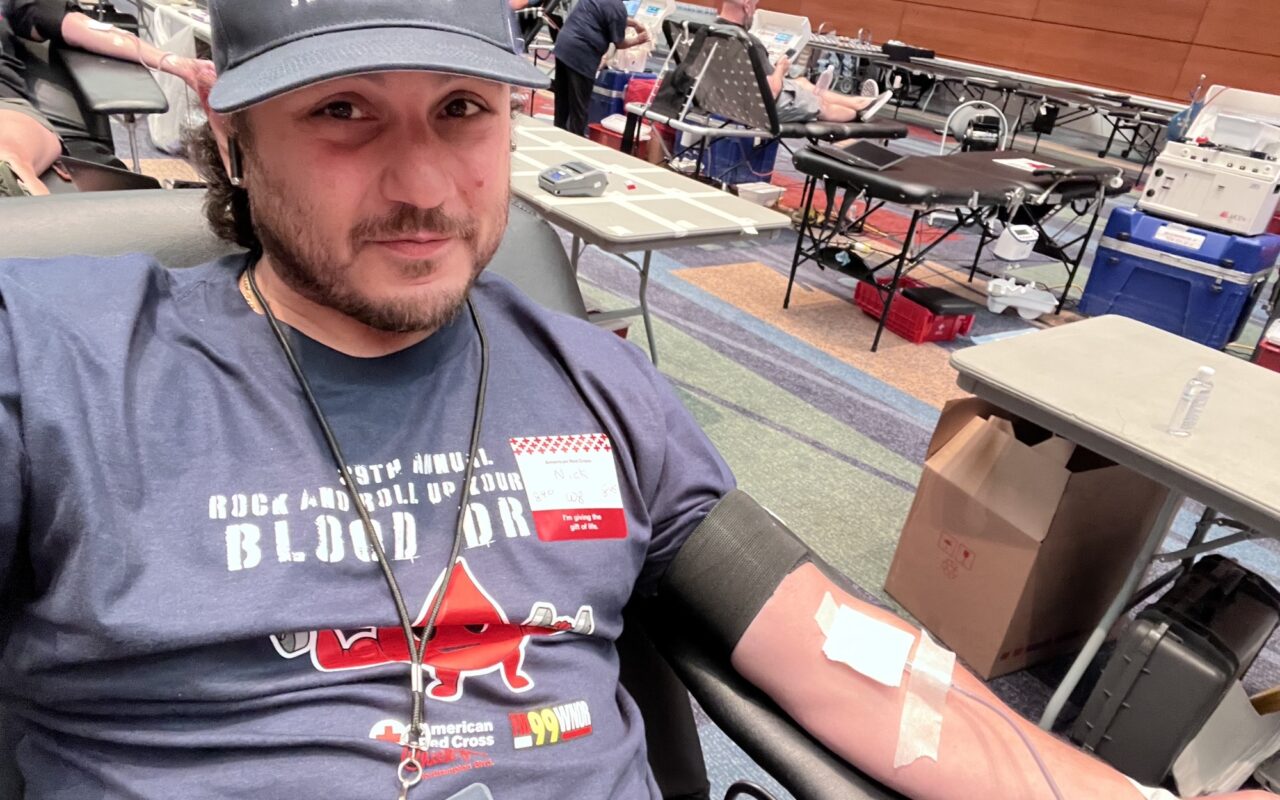 Man sitting in a donation chair, donating blood with a needle in his arm and bandage, at a blood drive center.