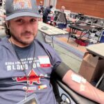 Man sitting in a donation chair, donating blood with a needle in his arm and bandage, at a blood drive center.