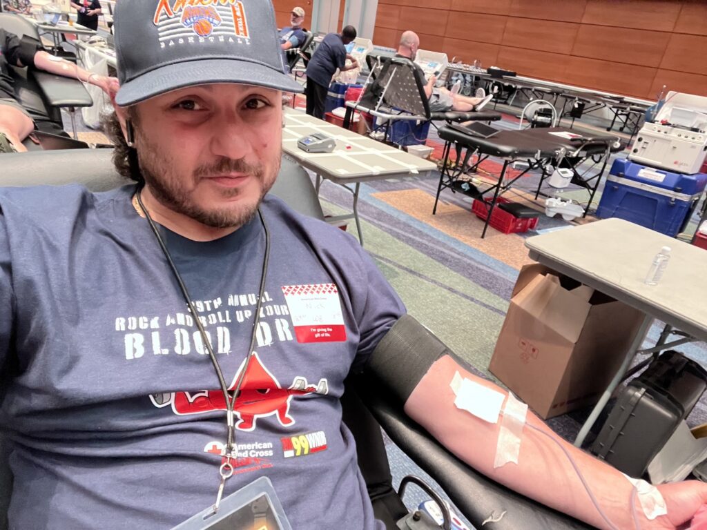 Man sitting in a donation chair, donating blood with a needle in his arm and bandage, at a blood drive center.