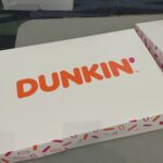 White Dunkin' box with an orange logo on a desk; a cardboard Gildan box and stacks of paper are visible in the background.