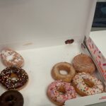 Assorted Dunkin' donuts in an open box on a table, including chocolate with sprinkles, pink frosted with sprinkles, glitter glaze, and powdered-sugar varieties.