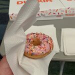 Donut with pink icing and rainbow sprinkles held in a white napkin, Dunkin boxes in the background.
