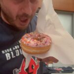 Man holding a pink-frosted donut with rainbow sprinkles on a napkin, in a kitchen setting.