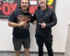 Two men pose indoors in front of radio station posters, smiling with thumbs up.